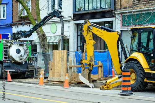 Photography vacuum pump truck and backhoe at work site on city street with shop fronts in ba