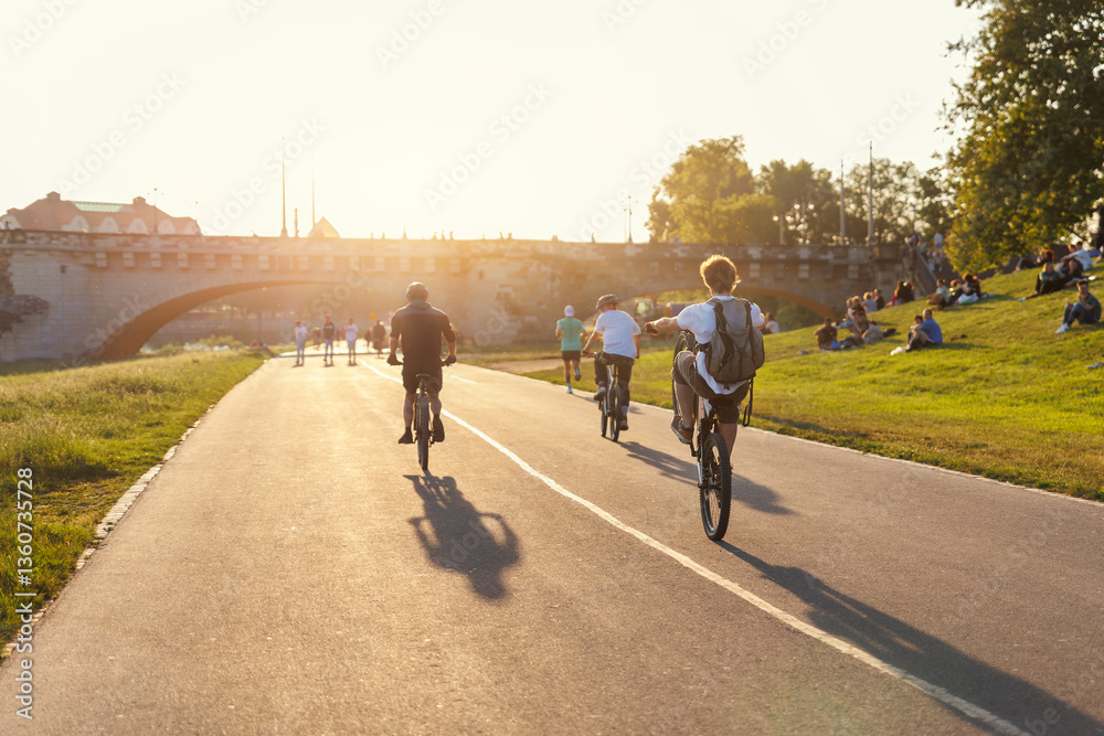 Obraz premium Scenic view youth people ride bicycles sunny Dresden Elbe riverside path against warm sunset glow active leisure. Rider perform wheelie beside historic Augustus bridge crowd relaxes grass lawn meadow