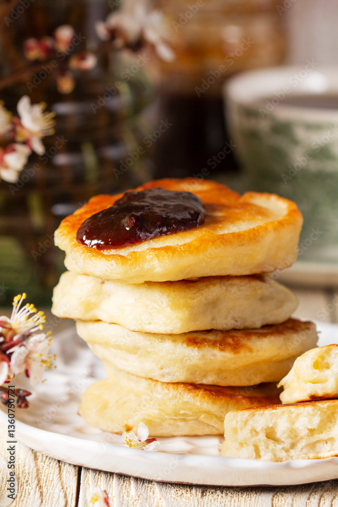 Lenten yeast pancakes with plum jam and tea, and a bouquet of flowering apricot branches on a light wooden table.
