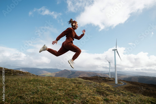 Young sportswoman jumping while listening music near wind turbines