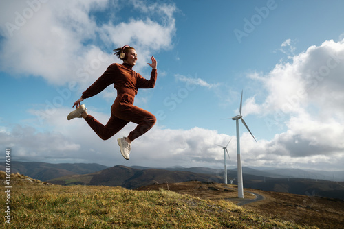 Young sportswoman jumping listening music with wind turbines and mountains in background