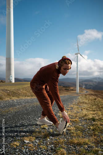 Young sportswoman stretching legs and listening music near wind turbines