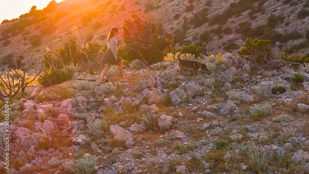 AERIAL, LENS FLARE: Young woman hikes through rocky hillside with her dog during golden hour. Surrounded by Mediterranean shrubs and glowing sunlight, they set off for morning walk in pristine nature.
