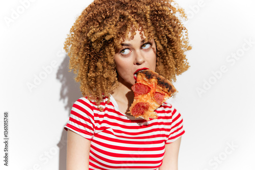 Young woman with curly hair in a striped shirt playfully biting a slice of pizza, conveying a fun and carefree attitude, perfect for food and lifestyle themes