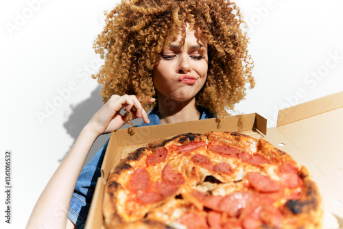 A disappointed woman with curly hair holds a pizza box with pepperoni pizza, showcasing her indecision and craving for unhealthy food in a bright, minimalist setting