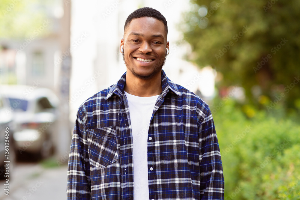 Fototapeta premium Portrait of happy black hipster wearing earbuds and posing at camera, spending time outdoor, selective focus