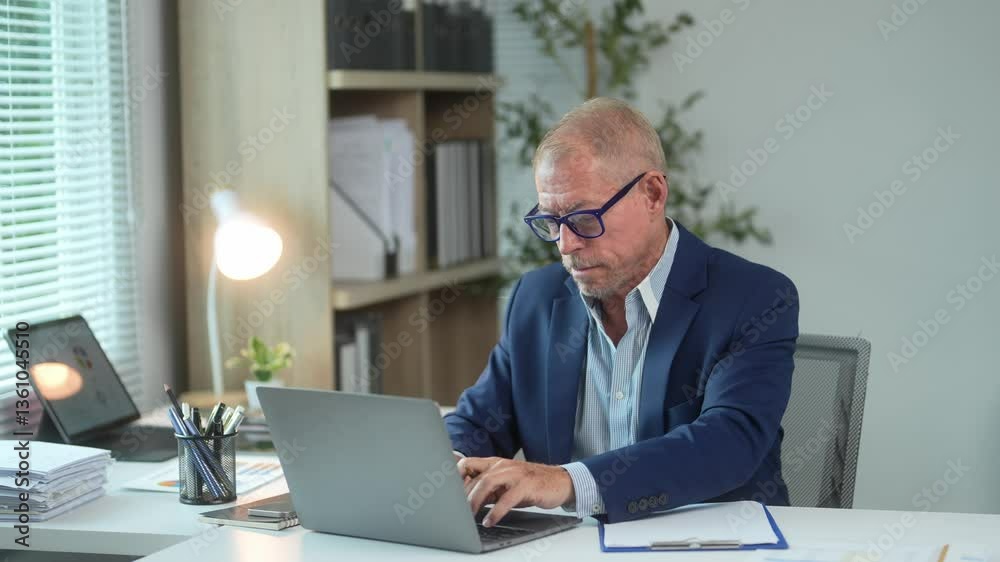 A man in a suit is sitting at a desk with a laptop in front of him. He is typing on the laptop and he is focused on his work A man in a suit is sitting at a desk with a laptop in front of him. He is typing on the laptop and he is focused on his work
