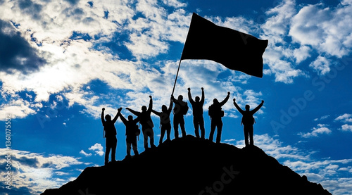 Silhouette group of people on mountain summit holding a flag against a blue sky