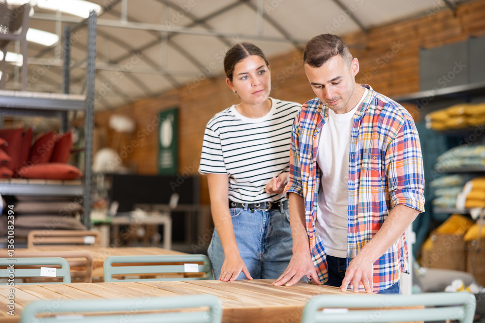 Obraz premium Positive young couple, girl and guy inspecting outdoor furniture at home and garden store, choosing patio table and enjoying time together