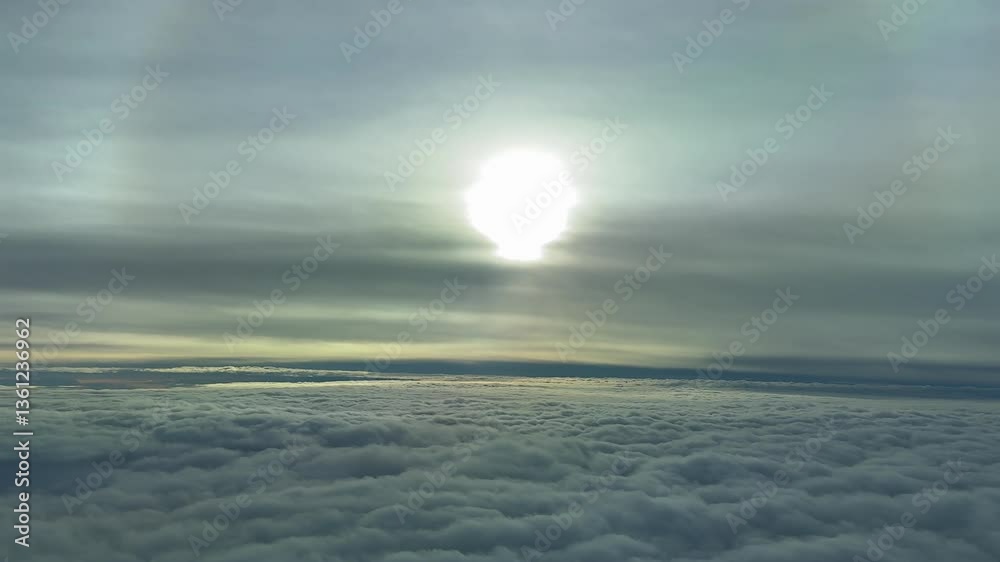 An piloit’s view from a jet cocpkit, recorded at sunrise while flying at the blue hour over a sea of clouds and a sun velied by fluffy clouds with an hallo.