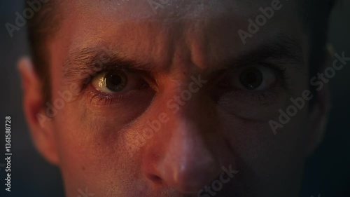 Close-up of angry man's eyes staring with anger at night illuminated by red light. Horror scene aggressive psychopath looking at camera. Macro shot angry male eyes close-up