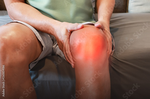 An elderly man sits on a sofa at home, holding his knee which is sore from osteoarthritis. Knee joint injury.