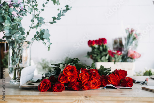 Red rose bouquet ready for packing in florist studio with eucalyptus in vase. Reflects monthly floral subscriptions and romantic gifting rituals