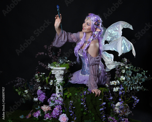 full length portrait of pretty female model wearing fantasy elf costume with fairy wings and purple dress. sitting pose on magical dark studio background with romantic mossy flowers.
