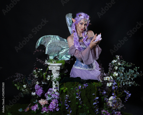 full length portrait of pretty female model wearing fantasy elf costume with fairy wings and purple dress. sitting pose, hands casting a magical spell,  magical dark studio background