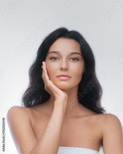 An elegant Asian woman with long black hair poses gracefully with her hand on her face in a skincare studio. Perfect for beauty, skincare, and cosmetic advertisements, emphasizing natural beauty and