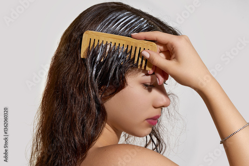 A young woman with long, dark brown hair is applying a hair treatment mask using a wide-tooth wooden comb.