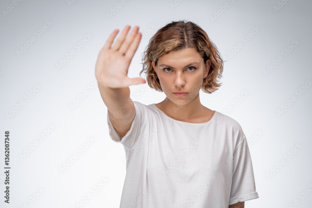 Fototapeta premium Young Caucasian woman in a white t-shirt extending her hand forward in a stop gesture with a serious expression. Conceptual image for refusal, rejection, boundaries, or protest themes.