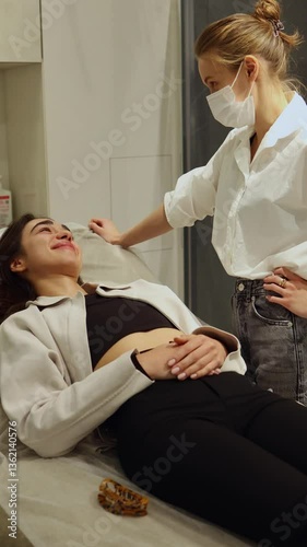 A young female patient lies on a treatment couch in a cosmetology clinic, while a doctor or cosmetologist consults her about skincare procedures