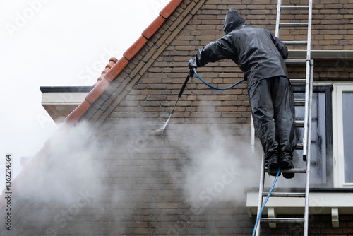 Man on ladder in waterproof suit cleans dirty brick facade of house with high pressure water jet. High pressure facade cleaning