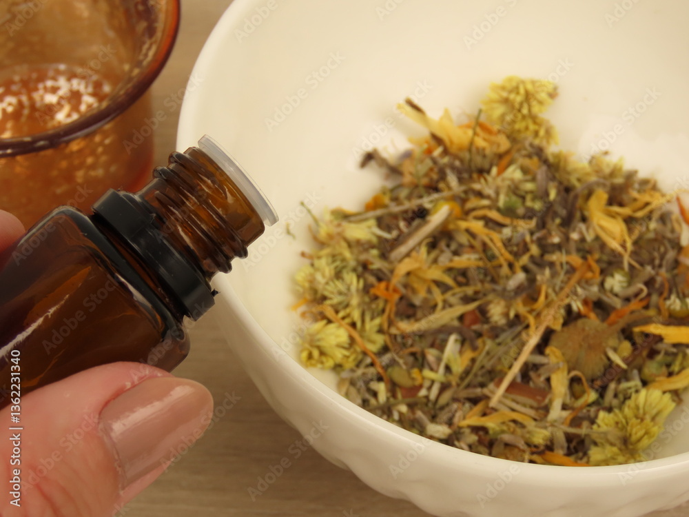 Fototapeta premium Woman hand holding a opened brown essential oil glass bottle over a white bowl with a mixture of dried herbs. Light wooden background. Self-care and wellness concept.