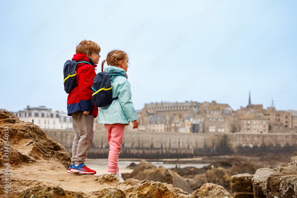 Two kids explore rocky coast with port town view in the distance