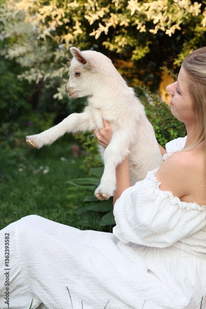 Fototapeta premium A young woman in a white dress sits on the grass in a garden, gently holding a white baby goat. The atmosphere exudes warmth and harmony with nature, with soft natural light creating a dreamy scene.