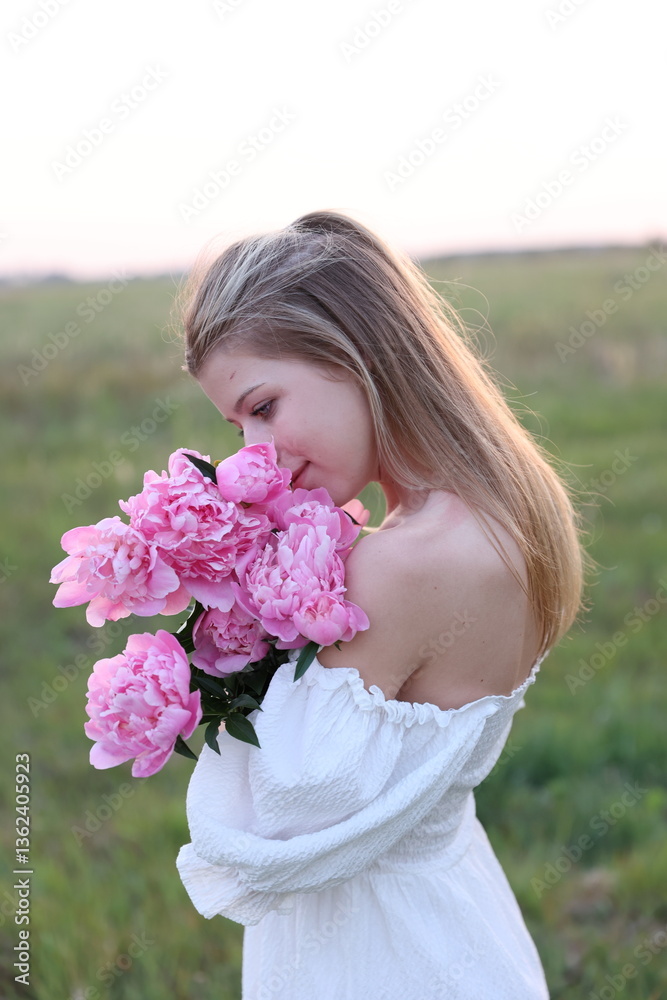Fototapeta premium A young woman in a white dress stands in a field at sunset, holding a bouquet of pink peonies. Her serene gaze and gentle expression convey a sense of peace and harmony with nature