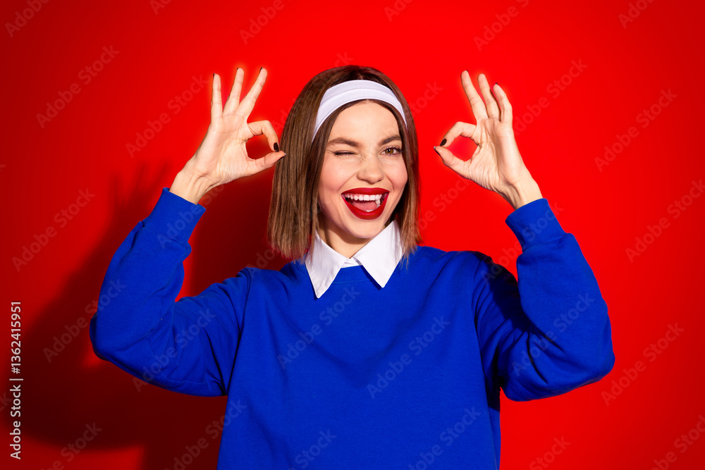Smiling Woman in Blue Sweatshirt Winking and Gesturing OK Sign on Bold Red Background Smiling Woman in Blue Sweatshirt Winking and Gesturing OK Sign on Bold Red Background