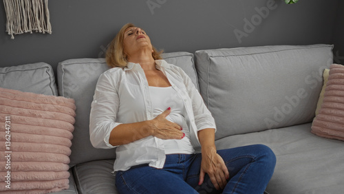 Elderly woman with stomach pain sitting on a sofa in a living room, looking uncomfortable in an indoor home setting.