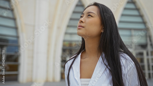 Free Download Young woman standing outdoors in an urban area looking up, with a modern city building in the background - GFXLoad