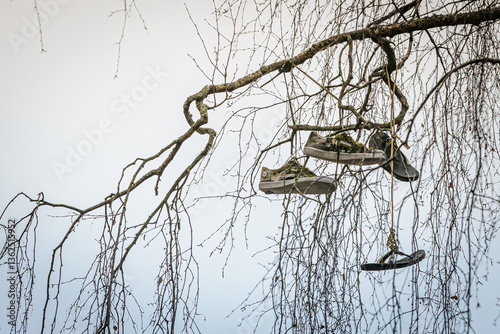 A pair of worn-out sneakers and a sandal hang from the bare branches of a tree against a pale sky, creating a moody and curious visual. Includes copy space.