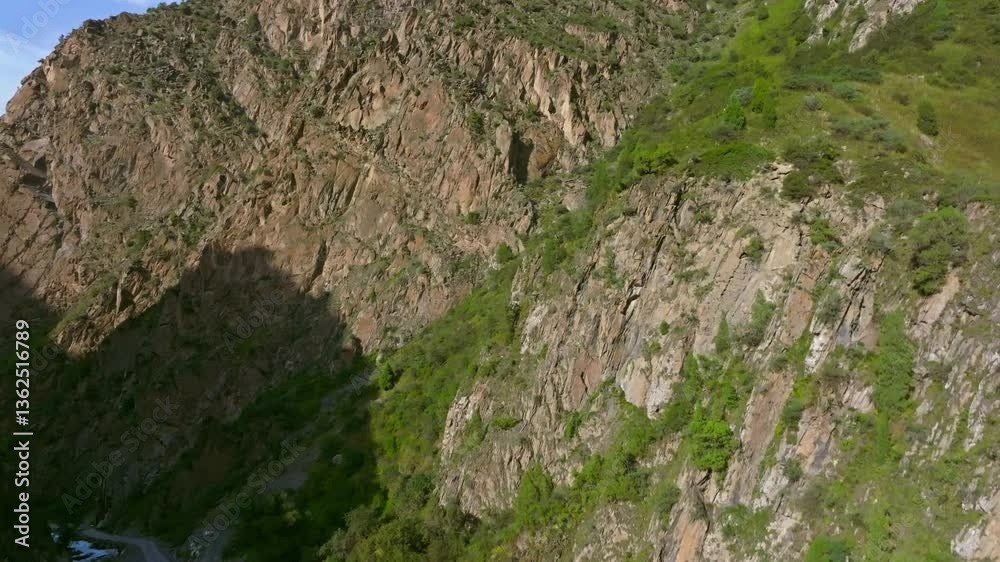 Aerial shot of a steep, rocky mountain cliff in Kyrgyzstan, descending into a lush green valley with a winding dirt road. Sunlit rocks contrast with shadowed areas, showcasing natural rugged beauty.