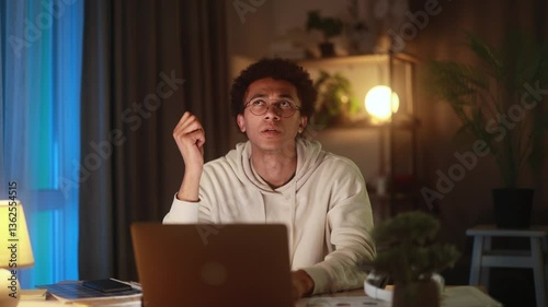 Young man sitting at laptop at home, suddenly getting new idea or insight, raising hand in realisation while working or thinking in cozy evening atmosphere, inspired by creative breakthrough