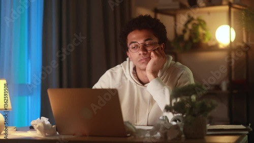 Exhausted young man resting head on hand while staring blankly at laptop, feeling mentally drained, unmotivated or bored while working or studying late at home in a quiet evening

