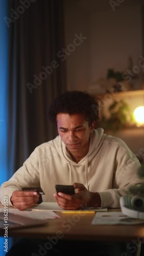 Vertical video of frustrated young African American man holding credit card and smartphone at home, experiencing online payment failure or banking issue while shopping or making transaction