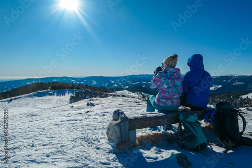 Obraz na płótnie Couple sits on a wooden bench enjoying sunny winter day atop snow-covered Wölkerkogel in Styria, Austria. Panoramic view of majestic snow-capped mountain peaks in Lavanttal Alps. Snowshoe hiking w salonie