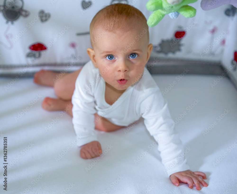 Fototapeta premium A 3-5 month old boy lying on his tummy on a white bed in a white bodysuit and looking at the camera. High quality photo