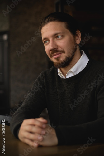 A man with his hair pulled back sits in a cafe. A man in a green sweater with long hair pulled back in a ponytail and a beard