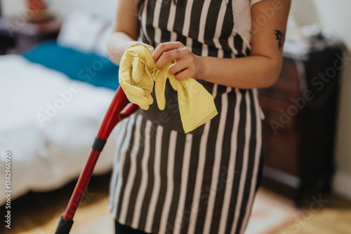 A lady wearing a striped apron and yellow gloves holds a broom handle, set in a cozy domestic interior, emphasizing household organization and cleanliness.