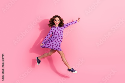 Delightful young girl in a stylish polka-dot dress jumps joyfully against a bright pink background