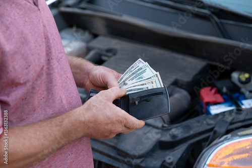 Man holds dollar bills while assessing car repairs with determination. A man examines his vehicle's engine with a wad of cash, contemplating repair costs and solutions outside his home
