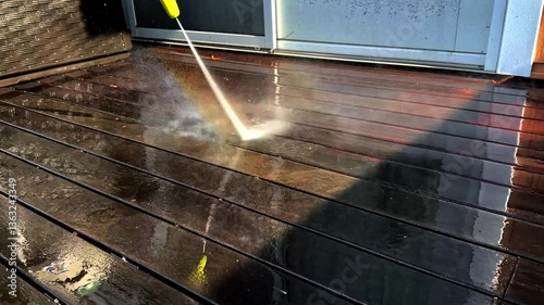 A pressure washer sprays water across a wooden deck, removing dirt and grime. Bright sunlight enhances the natural colors of the wood while droplets glisten on the surface
