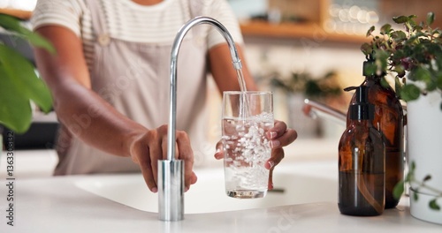 Hands, glass and water by faucet in kitchen for hydration, thirst or wellness in morning at apartment. Person, container and pouring liquid by sink with tap for drink, detox and gut health in house photo