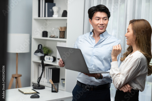 Smiling Asian coworkers standing while talking planning marketing report project on laptop casual day at modern office. Analyzing successful customer need in new social media online channel. Infobahn.