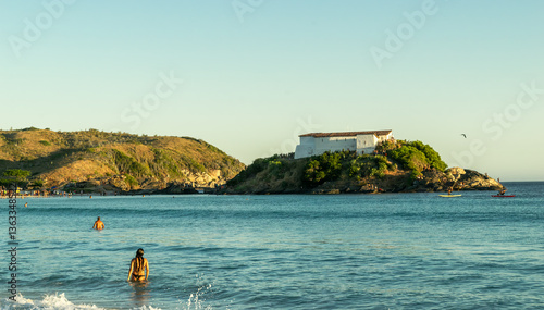 Back side at sunset, highlighting the last rays of sunlight illuminating the Praia do Forte fort and some bathers in the water. Cabo Frio, Rio de Janeiro, Brazil.