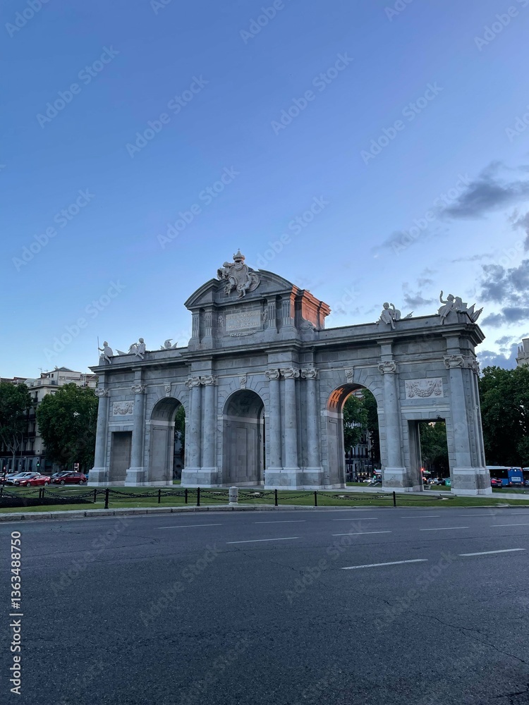 Obraz premium Stunning view of Puerta de Alcalá in Madrid at dusk with a serene blue sky. This neoclassical triumphal arch showcases intricate carvings and stands as an iconic landmark in Spain's vibrant capital.