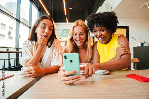 Fotografi Three joyful women excitedly sharing a delightful moment while watching something amusing on their smartphone in a trendy coffee shop