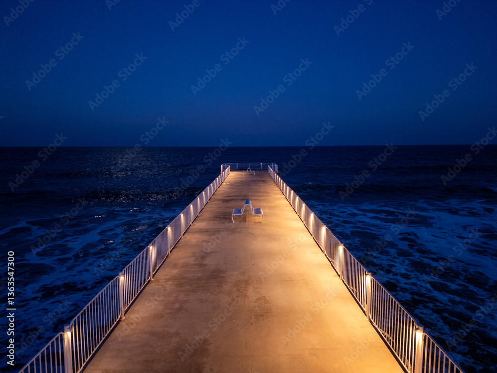 Obraz premium Long illuminated pier stretching into the dark sea at night, with calm waves and a deep blue sky, creating a peaceful and solitary coastal atmosphere