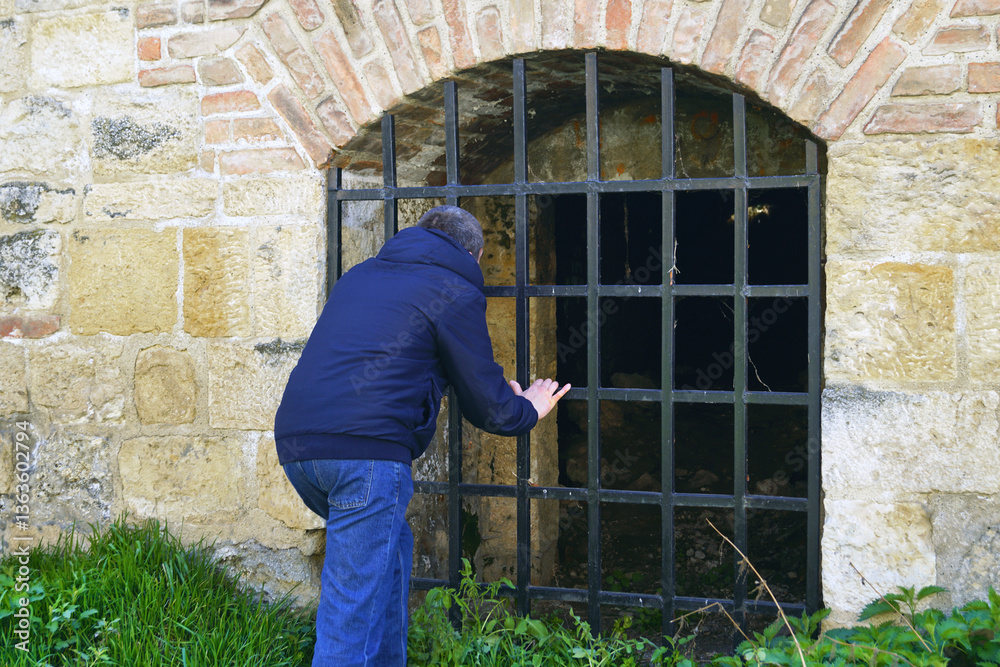 A man peers into the fortress's dungeon: a tourist leaning over the bars is trying to see from the outside the contents of a dark room built inside the defensive wall. Exploring abandoned buildings A man peers into the fortress's dungeon: a tourist leaning over the bars is trying to see from the outside the contents of a dark room built inside the defensive wall. Exploring abandoned buildings